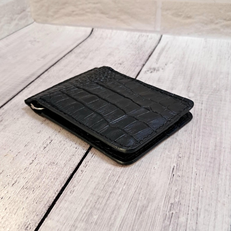 May include: A black leather wallet with a crocodile skin texture. The wallet has a metal money clip on the side. The wallet is rectangular and appears to be made of high-quality leather.