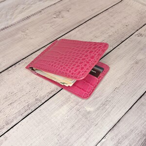 May include: A bright pink wallet with a crocodile skin effect. The wallet is open, showing banknotes and a debit card. The word "debit" is visible on the card. The wallet is made of a glossy material.