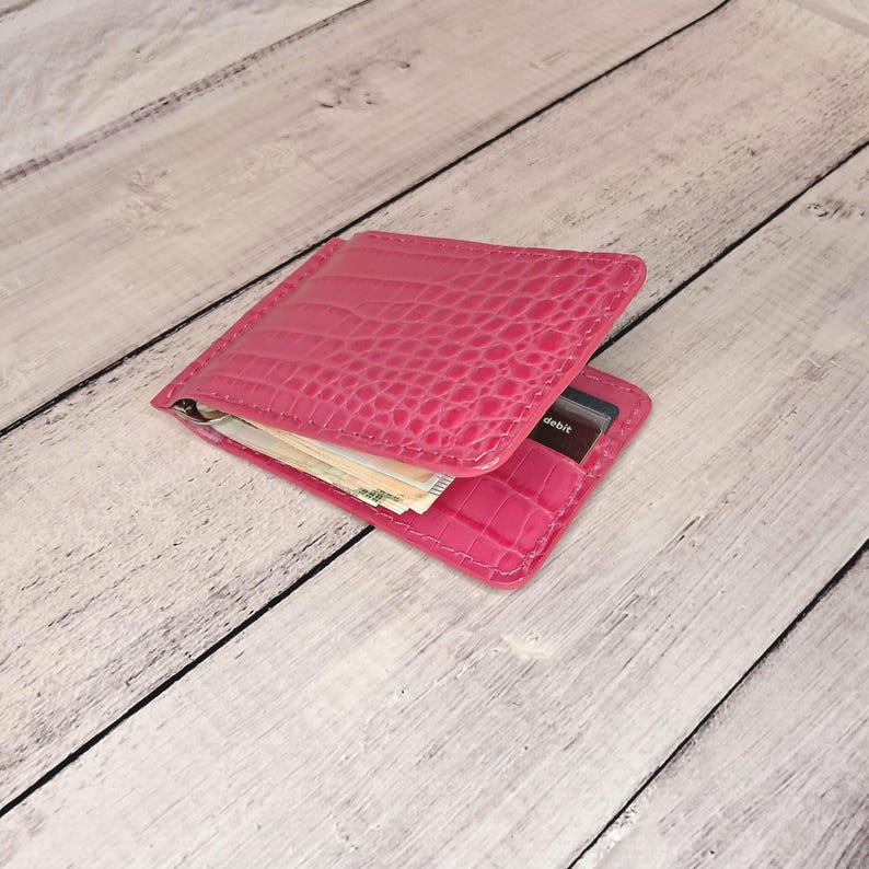 May include: A bright pink wallet with a crocodile skin effect. The wallet is open, showing banknotes and a debit card. The word "debit" is visible on the card. The wallet is made of a glossy material.