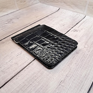 May include: A black wallet with a glossy, crocodile-effect finish. The rectangular wallet is closed and features a silver-coloured money clip. It rests on a light-coloured wooden surface.