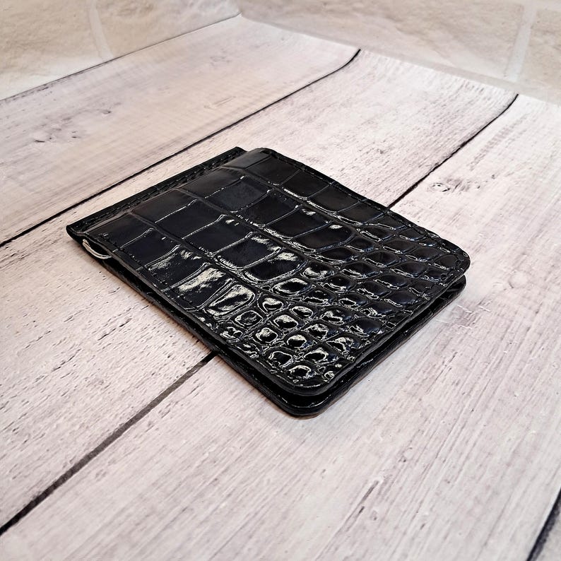 May include: A black wallet with a glossy, crocodile-effect finish. The rectangular wallet is closed and features a silver-coloured money clip. It rests on a light-coloured wooden surface.