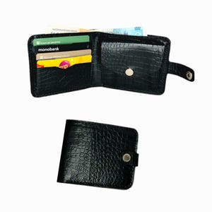 Puede incluir: Cartera de cuero negro con textura de piel de cocodrilo, abierta para mostrar ranuras para tarjetas y efectivo. La cartera se asegura con un cierre a presión. También se muestra una segunda cartera cerrada.