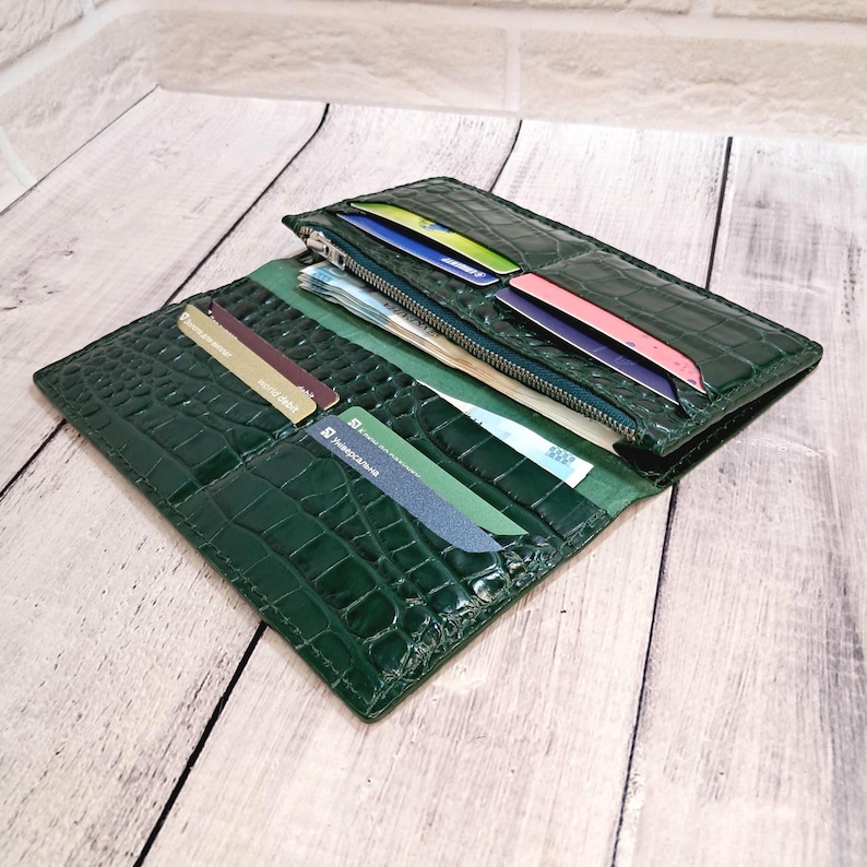Peut inclure: Un portefeuille vert fonc&eacute; avec un motif textur&eacute; en peau de crocodile. Le portefeuille est ouvert, r&eacute;v&eacute;lant plusieurs fentes int&eacute;rieures pour cartes contenant diverses cartes de cr&eacute;dit, un compartiment central zipp&eacute; et une section pour les billets.