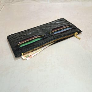 Könnte beinhalten: Eine schwarze, rechteckige Geldbörse mit Krokodillederstruktur. Die Geldbörse hat einen goldenen Reißverschluss und enthält mehrere Kreditkarten und gefaltete Geldscheine. Die Geldbörse ist geöffnet und zeigt den Inhalt.