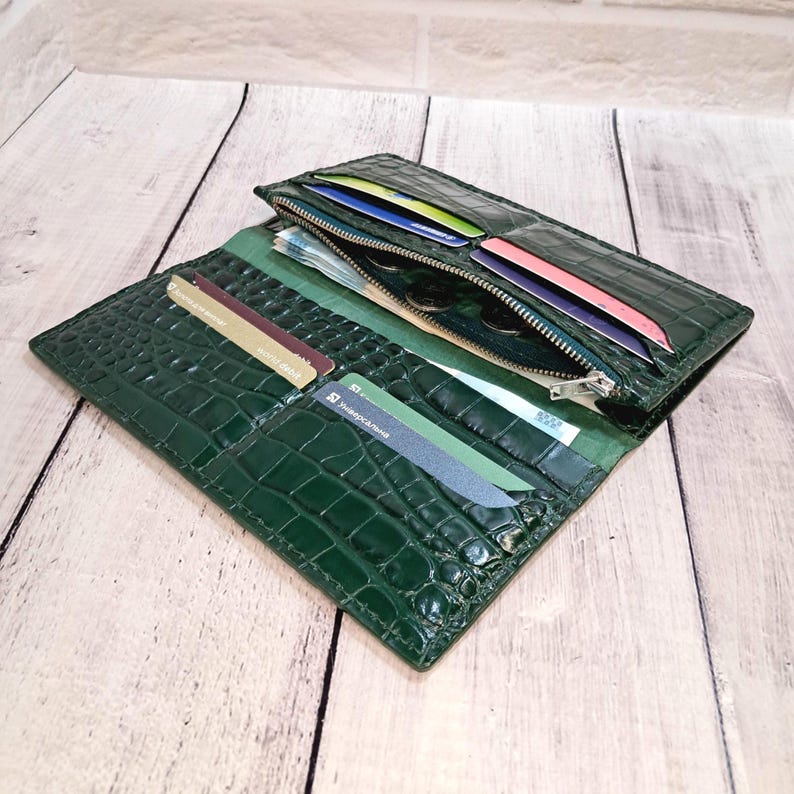 Peut inclure: Un portefeuille long vert fonc&eacute; avec un motif textur&eacute; en peau de crocodile. Le portefeuille ouvert pr&eacute;sente plusieurs fentes pour cartes, un compartiment central zipp&eacute; et plusieurs poches pour billets contenant diverses cartes et billets.