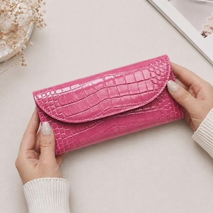 Peut inclure: Un portefeuille rectangulaire rose vif doté d'une finition brillante et d'une texture en relief façon peau de crocodile. Le portefeuille présente un rabat incurvé qui recouvre le panneau avant, affichant un motif d'écailles embossé uniforme.