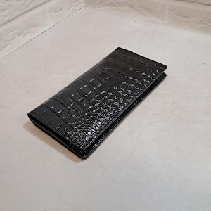 Peut inclure: Un portefeuille rectangulaire noir dot&eacute; d'une finition brillante et d'un motif textur&eacute; en peau de crocodile. Le portefeuille est pli&eacute; et ferm&eacute;, pr&eacute;sentant un motif d'&eacute;cailles gaufr&eacute; coh&eacute;rent sur toute la surface ext&eacute;rieure de l'objet.