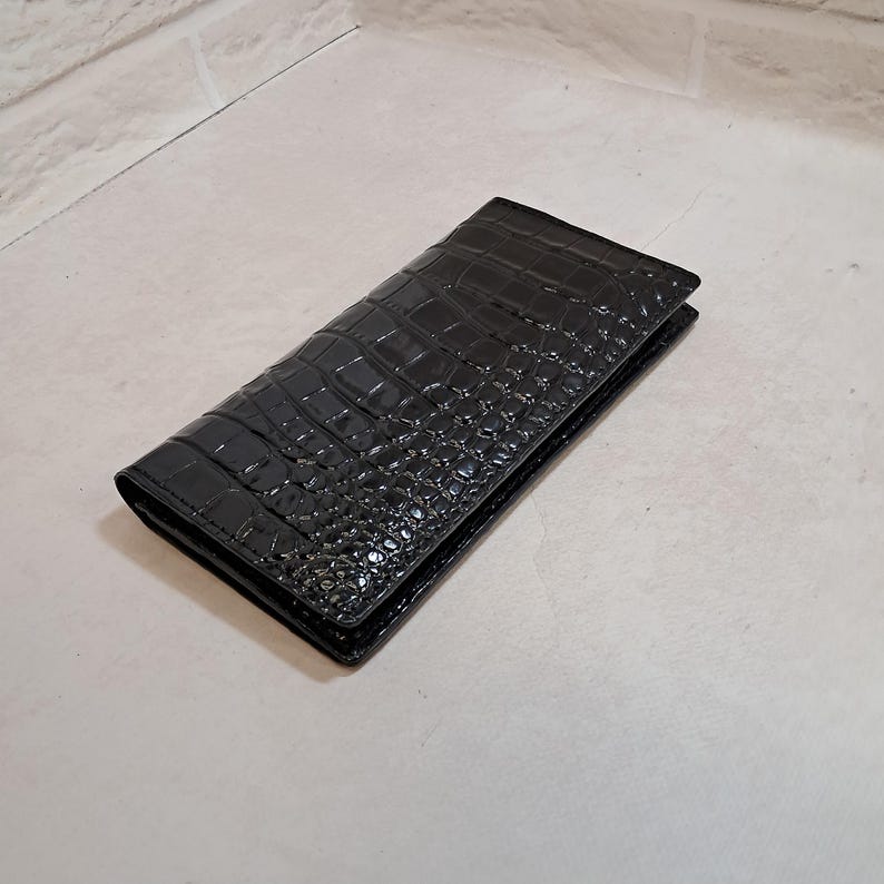 Peut inclure: Un portefeuille rectangulaire noir dot&eacute; d'une finition brillante et d'un motif textur&eacute; en peau de crocodile. Le portefeuille est pli&eacute; et ferm&eacute;, pr&eacute;sentant un motif d'&eacute;cailles gaufr&eacute; coh&eacute;rent sur toute la surface ext&eacute;rieure de l'objet.