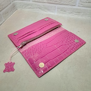 Könnte beinhalten: Offenes, leuchtend pinkes Portemonnaie mit Krokodilleder-Optik. Es hat einen Reißverschluss, mehrere Kartenfächer und einen Druckknopfverschluss. Ein kleines, pinkfarbenes Lederetikett ist angebracht.