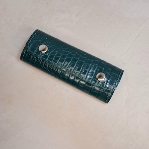 Puede incluir: Un estuche para llaves de color verde azulado con textura de piel de cocodrilo sintética. El estuche rectangular se cierra con dos broches plateados. El estuche está enrollado y parece vacío.