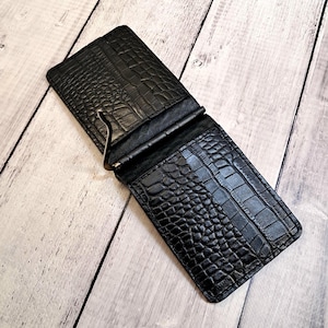 May include: Black leather money clip wallet with a crocodile skin texture. The wallet is open, revealing a metal money clip. The wallet is rectangular and has a textured surface. The wallet is on a light wood surface.