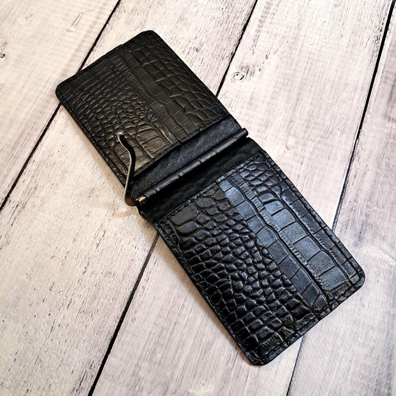 May include: Black leather money clip wallet with a crocodile skin texture. The wallet is open, revealing a metal money clip. The wallet is rectangular and has a textured surface. The wallet is on a light wood surface.