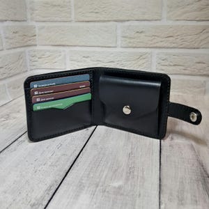 Peut inclure: Portefeuille en cuir noir ouvert, avec plusieurs fentes pour cartes et une petite poche à monnaie. Le portefeuille est ouvert, révélant plusieurs cartes de crédit colorées. Une fermeture à bouton-pression argentée sécurise la poche à monnaie.