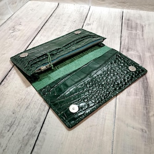 May include: Open, dark green leather wallet with a crocodile skin texture. The wallet has a zipper closure and multiple compartments. Silver snap closures secure the wallet. The interior is a lighter shade of green.