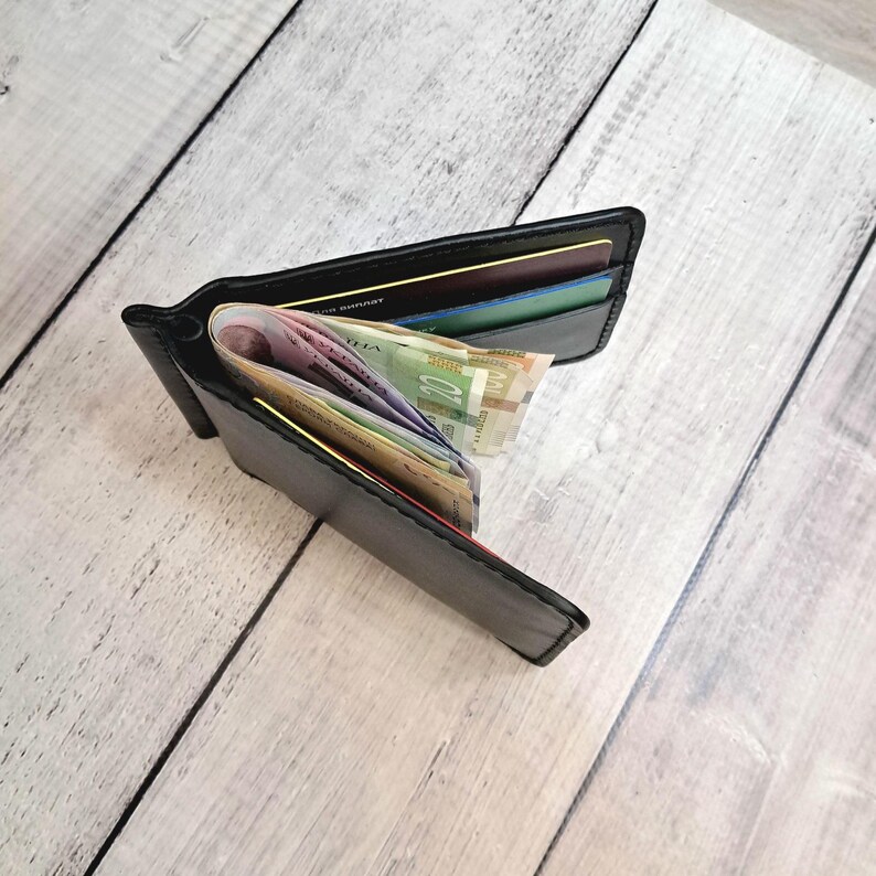 May include: Open black leather wallet filled with colourful banknotes and credit cards. The wallet is displayed open, showcasing the contents. The background is a light-coloured wooden surface.