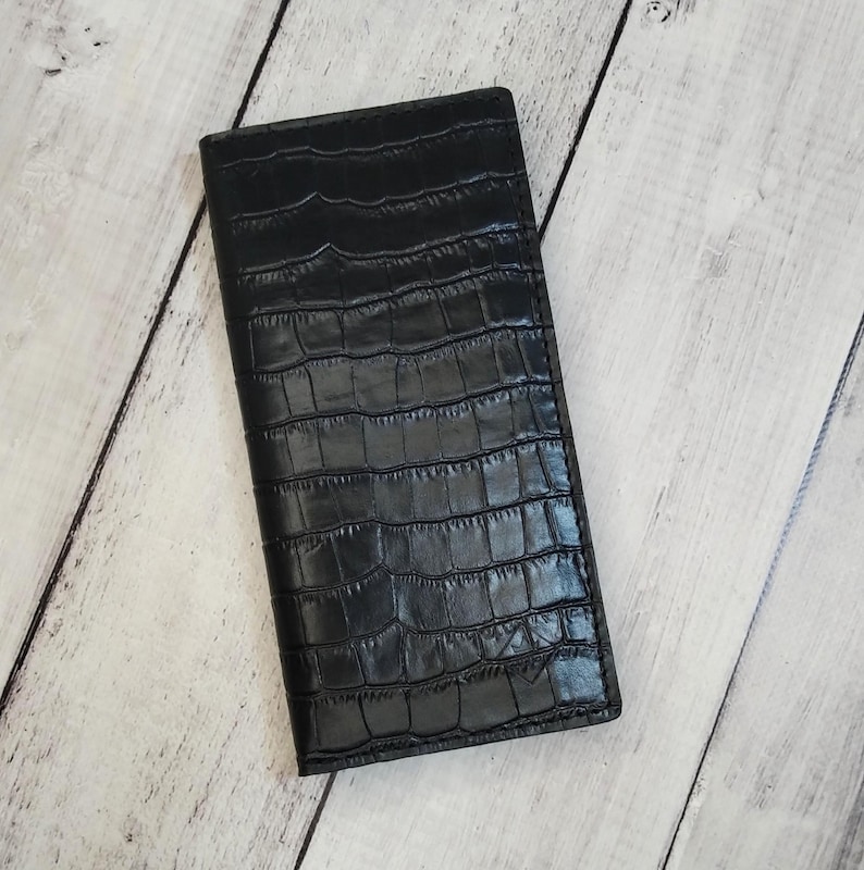 Peut inclure: Un portefeuille rectangulaire noir pr&eacute;sentant un motif textur&eacute; en peau de crocodile sur toute la surface. Le portefeuille a un design fin et allong&eacute; avec des coutures visibles le long des bords et une finition lisse et uniforme sur l'ext&eacute;rieur.