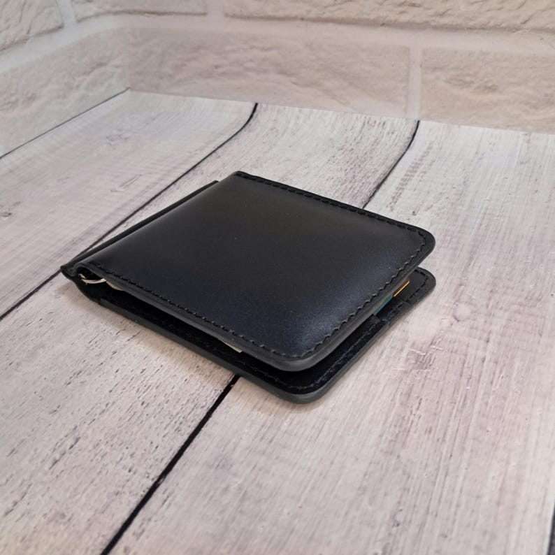 May include: A black leather money clip wallet. The wallet is rectangular with rounded corners and a silver-coloured clip. The edges are stitched with black thread. The wallet is open, revealing a glimpse of the interior.