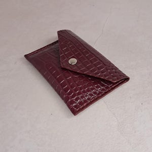 Può includere: Un portafoglio bordeaux in similpelle con una trama a pelle di coccodrillo. Il portafoglio ha una chiusura a patta assicurata da un bottone a pressione argentato. Il portafoglio è rettangolare e sembra vuoto.