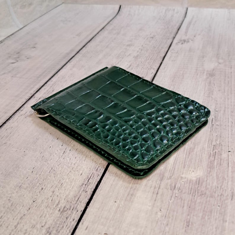 May include: A dark green leather wallet with a crocodile skin effect. The rectangular wallet has a silver money clip. The wallet is closed and rests on a light wood surface.