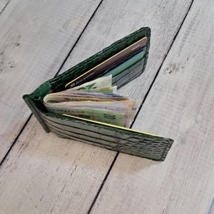 May include: Open green leather wallet with a crocodile skin texture, filled with cash and credit cards. The wallet is open, displaying the contents. The background is a light-coloured wooden surface.
