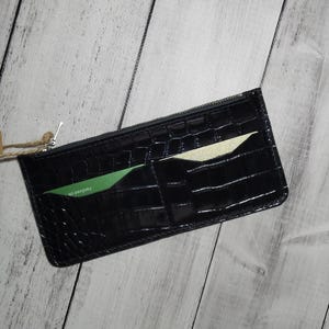 Puede incluir: Cartera negra texturizada con cierre de cremallera. La cartera tiene ranuras para tarjetas con una tarjeta verde y una crema visibles. Una pequeña etiqueta con texto cuelga de la cremallera. La cartera está sobre una superficie de madera clara.