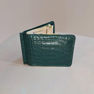 May include: A dark green bifold money clip wallet featuring a textured crocodile skin pattern and matching green stitching. The wallet is partially open, revealing a stack of paper currency with the text UKRAINA printed on the visible banknote.