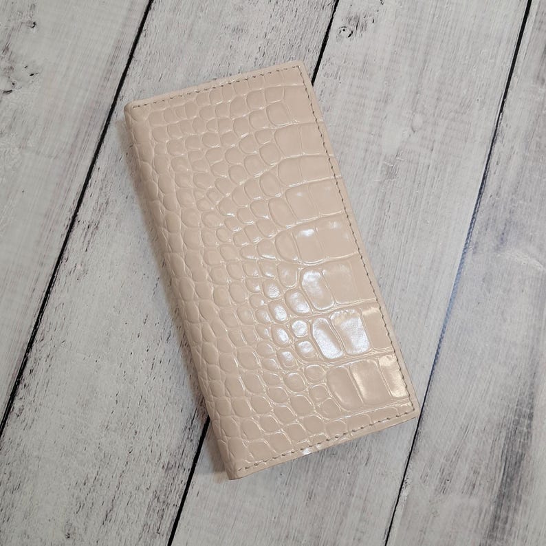 Peut inclure: Un portefeuille rectangulaire beige clair dot&eacute; d'une texture gaufr&eacute;e brillante fa&ccedil;on peau de crocodile. Le portefeuille pr&eacute;sente un profil fin et allong&eacute; avec des bords cousus nets sur tout le p&eacute;rim&egrave;tre, affichant un motif reptilien.