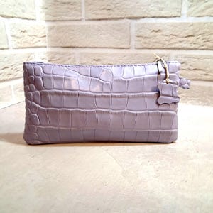 May include: A lavender-colored, rectangular clutch with a crocodile skin texture. The zipper is black, and a small leather tag hangs from the side. The bag is sitting on a light-colored surface in front of a brick wall.