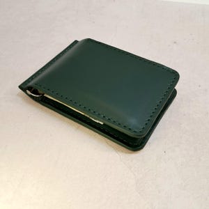 Könnte beinhalten: Eine dunkelwaldgrüne Geldklammer-Geldbörse aus Leder mit glatter, matter Oberfläche und farblich abgestimmten Nähten an den Kanten. Das rechteckige Accessoire hat ein schmales, gefaltetes Design mit einem sichtbaren Metallclip an der Seite.