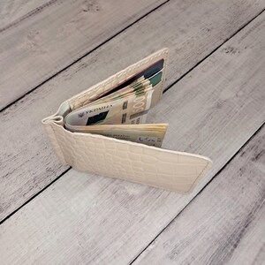 May include: Open, pale beige wallet with a textured, crocodile-effect pattern. The wallet is filled with folded banknotes and credit cards. The wallet is open, displaying the contents.