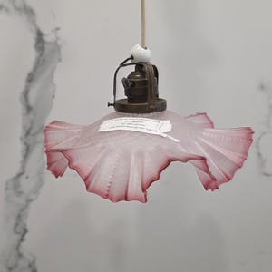 Könnte beinhalten: Vintage Hängeleuchte mit einem gerüschten, rosa Glasschirm. Der Schirm hat einen dunkelrosa Rand und hängt an einer Messing-farbenen Metallhalterung. Ein weißes Keramikelement und ein Kabel vervollständigen das Design. Hintergrund: eine weiß marmorierte Wand.