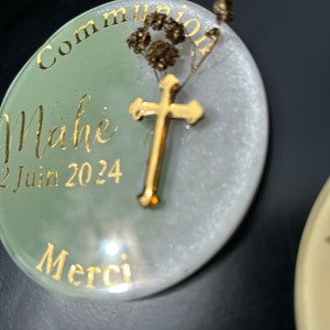 Puede incluir: Un recuerdo redondo de resina, blanco y verde, con letras doradas. El texto dice "Communion Mahé 22 Juin 2024 Merci". Una pequeña cruz dorada y flores secas están encima de la resina.