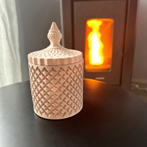 Peut inclure: Un pot de bougie en céramique rose avec un motif en diamant et un couvercle décoratif. Le pot est posé sur une surface noire devant une cheminée.