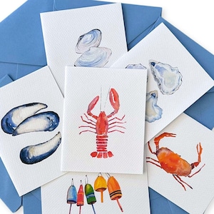 May include: Set of six watercolor greeting cards featuring various nautical themes. The cards feature illustrations of a lobster, a crab, a clam, an oyster, mussels, and buoys. The cards are printed on white cardstock and have a blue envelope.