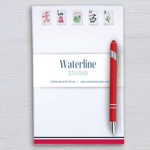 May include: A white notepad featuring five mahjong tile illustrations at the top, a central paper band labeled Waterline STUDIO, and a red ballpoint pen attached to the side. The bottom edge of the notepad has a horizontal red and black stripe.