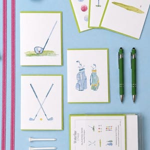 May include: A collection of golf-themed stationery and accessories on a light blue surface. The set includes watercolor-style greeting cards with golf course illustrations, two green pens, a golf ball, and golf tees. A pair of white golf gloves and pink striped ribbon are also present.