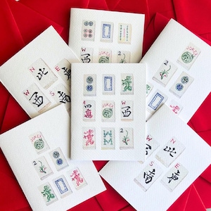 May include: White greeting cards featuring colorful illustrations of Mahjong tiles. The cards display various tile designs, including characters, dots, and symbols. The cards are arranged on a red background.