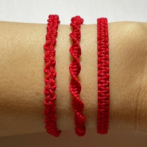 May include: Three red string bracelets with different knot patterns. The bracelets are worn on a person's wrist.