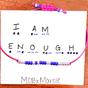 Op de afbeelding: Een roze koordarmband met blauwe en zilveren kralen in morsecode, gepresenteerd op een witte kaart met de tekst I AM ENOUGH, de bijbehorende morsecode-punten en streepjes, en de merknaam MCBxMarcie onderaan de kaart gedrukt.