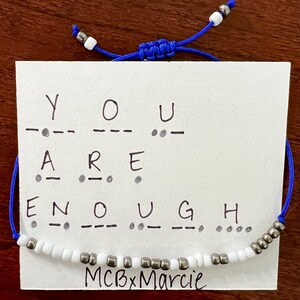 Op de afbeelding: Een blauwe koordarmband met witte en metallic kralen, gepresenteerd op een witte kaart met de tekst YOU ARE ENOUGH in zwarte inkt, inclusief bijbehorende morsecode-punten en streepjes. De kaart bevat onderaan de tekst MCBxMarcie.