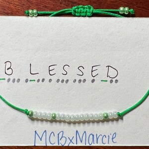 May include: A green cord bracelet featuring a row of small white and light green beads. The bracelet is displayed on a white card with the word BLESSED written in black ink above its corresponding Morse code dots and dashes, and MCBxMarcie below.
