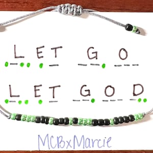May include: A gray cord bracelet featuring a row of alternating black and light green beads. The bracelet is displayed on a white card with the text LET GO and LET GOD written in black ink, accompanied by corresponding green Morse code dots and dashes.