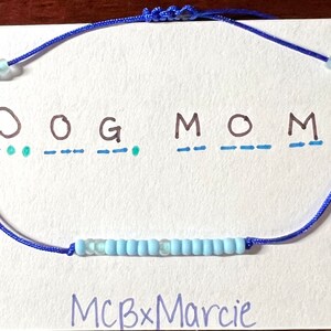 May include: A blue cord bracelet featuring a row of light blue beads, displayed on a white card with the text "DOG MOM" written in black ink above Morse code dots and dashes, and the text "MCBxMarcie" printed at the bottom of the card.