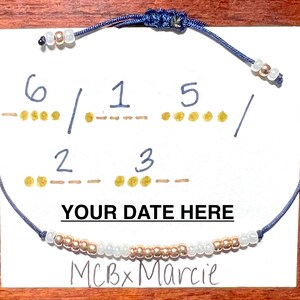 Puede incluir: Una pulsera de cordón azul con cuentas alternas en oro rosa y blanco, con un cierre de nudo deslizante ajustable. La pulsera se muestra sobre una tarjeta blanca con el texto "YOUR DATE HERE" y "MCBxMarcie" junto a código Morse dibujado a mano.