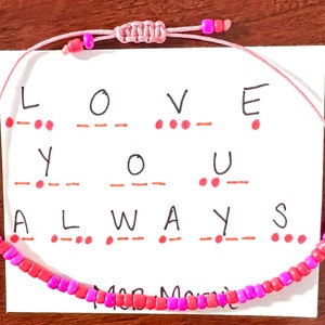 May include: A pink cord bracelet featuring alternating pink and coral beads, with an adjustable sliding knot closure. The bracelet is displayed on a white card printed with the words LOVE YOU ALWAYS and corresponding red Morse code dots and dashes.