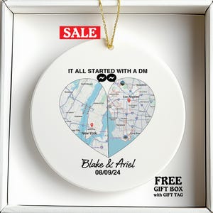 May include: White ceramic ornament with a map design split into two heart-shaped sections, featuring New York and Los Angeles. The text "IT ALL STARTED WITH A DM" is above the map, with names and a date below. A gold string is attached.