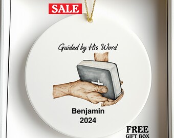 May include: White ceramic ornament with a gold string for hanging. The ornament features an illustration of a hand holding a book with a cross, and the text "Guided by His Word" and "Benjamin 2024". A red "SALE" tag is visible.