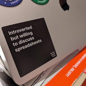 Op de afbeelding: Een zwarte sticker op een zilveren laptop met de tekst "Introvert maar bereid om spreadsheets te bespreken". De laptop heeft een Apple-logo. Andere stickers en een oranje doos met tekst zijn ook zichtbaar.