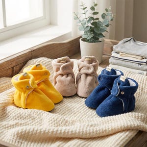 Può includere: Una collezione di scarpine per neonati gialle, beige e blu navy, disposte su una coperta color crema. Ogni paio presenta una chiusura con bottone e una piccola etichetta con la scritta "pufien". Le scarpine sono morbide e realizzate in un materiale felpato.