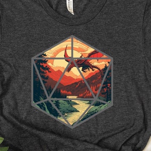 Puede incluir: Una camiseta gris oscuro con un gráfico de un dragón rojo volando sobre una cordillera dentro de una forma geométrica gris que se asemeja a un dado de veinte caras.